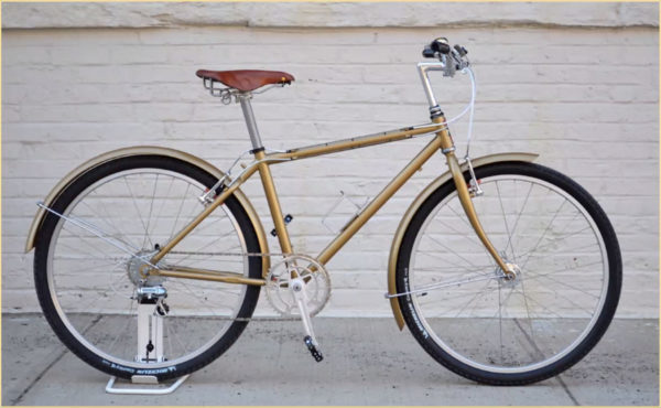 Restored gold bike with leather seat on the sidewalk with a white painted brick wall in the background.