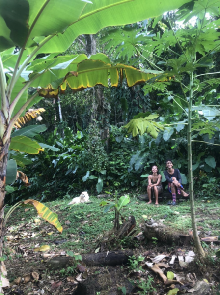 Alt Text: Rheanna and Gesiye sitting on a log in the forest. Their hiking dog, Grimey, with white fur sitting besides them. The forest is green from the vines, banana leaves and grass. 
