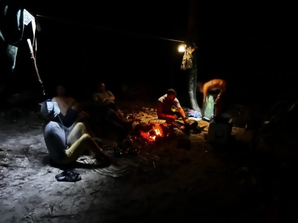  Alt Text: A group sitting around the bonfire at night. There are red embers from the fire, and camping lights hanging from above.