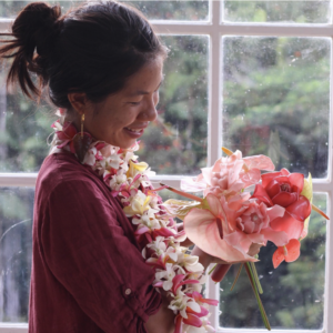 An image of a woman with dark hair in a bun, with a floral necklace around her neck. Wearing a pink shirt and holding a big bouquet of flowers. 