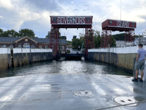 View standing from the front of a ferry with a wet, gray floor looking out Governors Island dock. There is water in the center with wooden walls with white paint at the top and a red and white sign overhead that reads “Governors Island.” Brick buildings are in the backdrop.