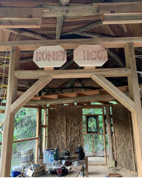 The doorway to the tiny house has wooden posts. There is the sign saying “Honey Hex” in front. Inside you can see walls constructed out of vetiver grass and clay. There are frames for windows to be attached. You can see the construction tools and materials for further building.