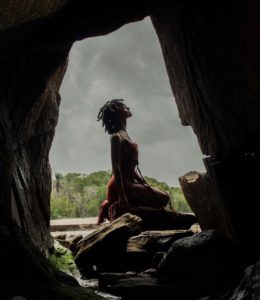 View of Gesiye perched on a rock, framed by a larger rock. She is wearing a long red dress, with her hair in short braids. The sky is overcast and grey. You see her side profile with eyes closed.