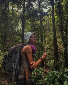 Tracy standing with a backpack, a cutlass/ machete by her side and a walking stick with a backdrop of the trees in the rainforest. She is wearing a light blue hat and glasses, and see her side profile.