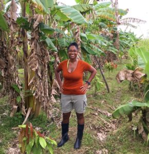 Aleya is wearing black boots and standing with hands on hips as she smiles in the field full of banana and plantain trees. There are young cacao saplings with green and red leaves growing in between.