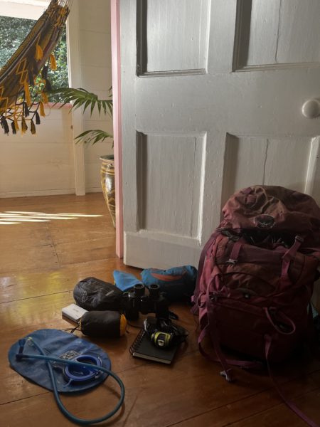 A pink backpack next to essentials like a device for drinking water, notebook, headlamp, rain pants, hammock and waterproof covering in case of rain 