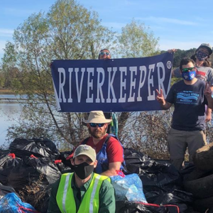 A group of people, one holding a sign saying "Riverkeeper", posed beside garbage bags in front of a river.