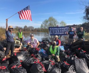 A group of people pose behind a large pile of black trash bags. One person holds an American flag aloft. Another person holds a banner with the word "Riverkeeper". A river is visible in the background.
