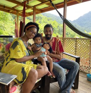 The family of 3 sit on a couch with the hills and vegetation in the background. You can see part of the lattice gate and hammock. Aleya is wearing a yellow dress, Michael wears an African textile short with jeans while their baby holds a stuffed toy
