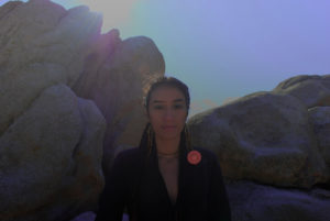 Image of Sarah in front of a rock structure. A person with dark hair and braids, the sun flares over their face. 