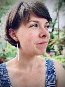 An image of a white person with a short haircut, nose ring, smiling with their face turned toward the right. The background is a blurry garden. 
