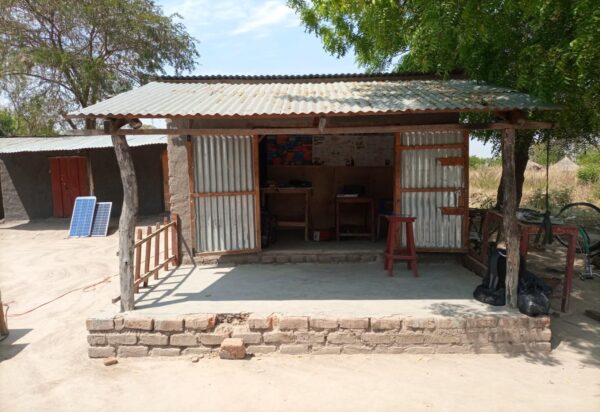 Front view of CC4D Bright Makerspace in Eden II Village, Rhino Camp Refugee Settlement, a single unit building structure with open metal doors, a brick front, wood table and stools, with trees in the background.