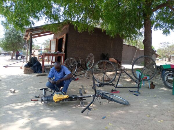 Volunteer Mr. Kigimingi is kneeling down and repairing a bicycle laid out on the ground at the Bright Makerspace in 2022, with two other bicycles standing up in the background.