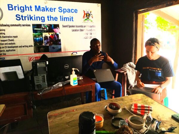 Mathew sitting down with a laptop on his lap, briefing EU volunteer Ambra Malandrin who is also sitting with a laptop on her lap. There are two desks, one with laptops, and another with repair materials such as tape and other devices, and a poster in the background with the title “Bright Maker Space Striking the Limit”