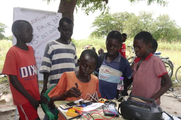A group of five children, four standing up and one seated at a table holding a repair tool, learning how to repair a solar lamp during the 2022 International Repair Day Celebration public event in Eden Village, Rhino Camp Refugee Settlem