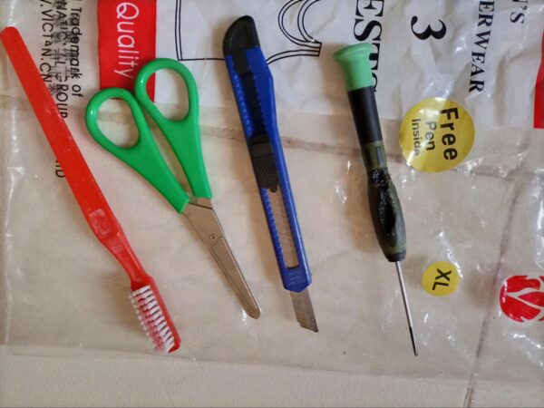 A red toothbrush, green scissors, a cutter knife, and a screwdriver over a zip lock plastic bag. 