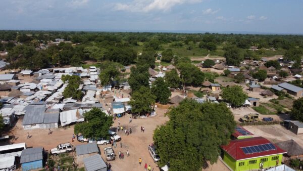 An aerial view of the Ocea Village with most shelters build with muddied walls and grass touched roofing in Rhino Camp Refugee Settlement, Arua - Uganda - the image depicts housing structures, trees, and a partially-cloudy sky.