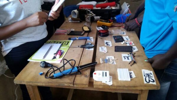 Three people that are part of an Open Tech training are by a wooden table that contains soldering iron, LED tester, breadboard, censor, and registers.
