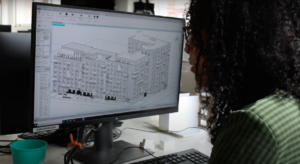 A black woman, facing away from the camera, sits in front of a computer screen and edits a building modeled on 3D software.