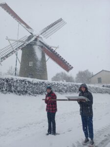 Two students in the Netherlands stand on a snowy grey day beside a windmill