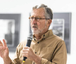 An image of a white man in a tan shirt holding a microphone.