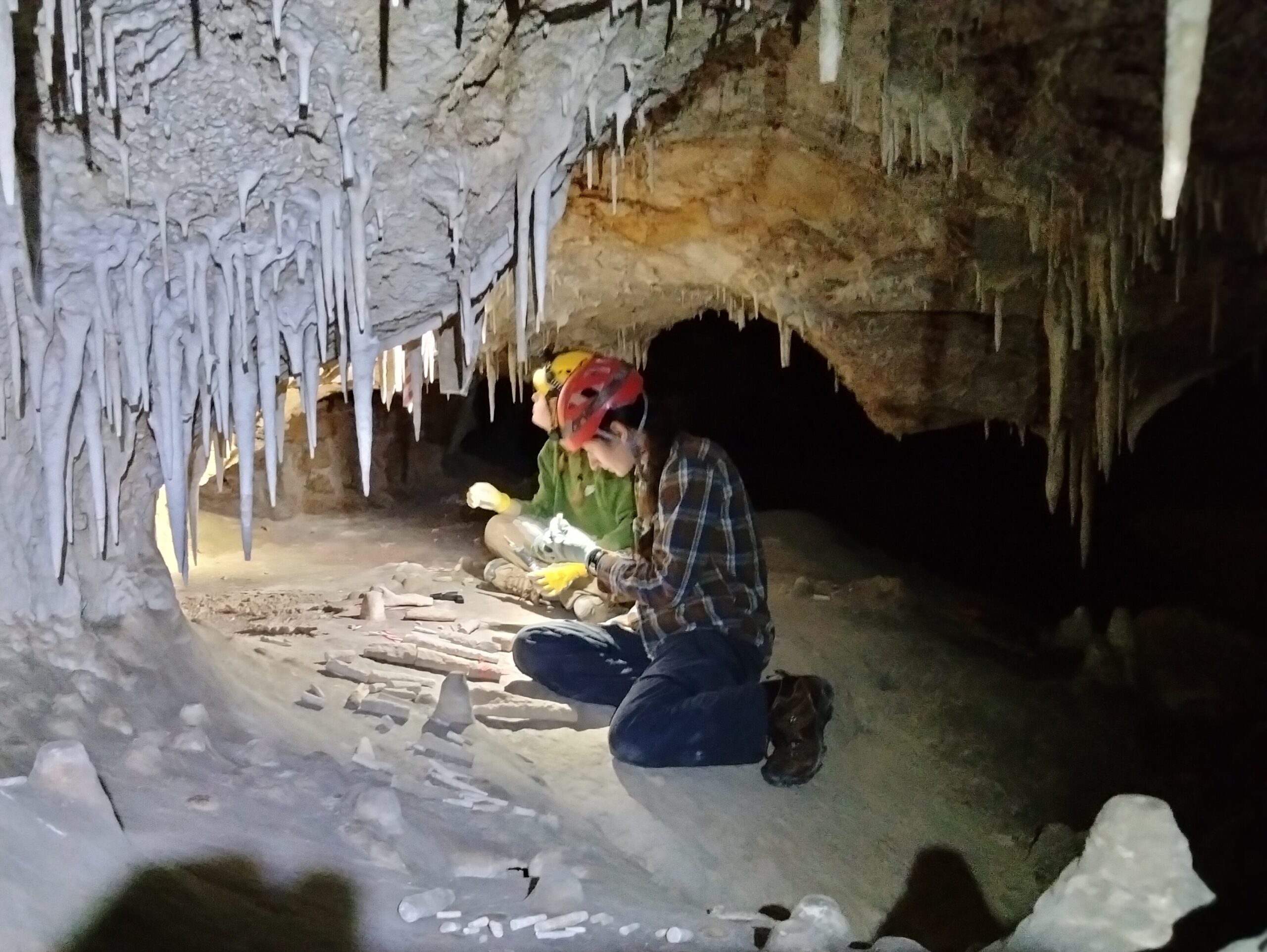 Two people sitting in the floor of a cave, look closely into a group of broken speleothems.