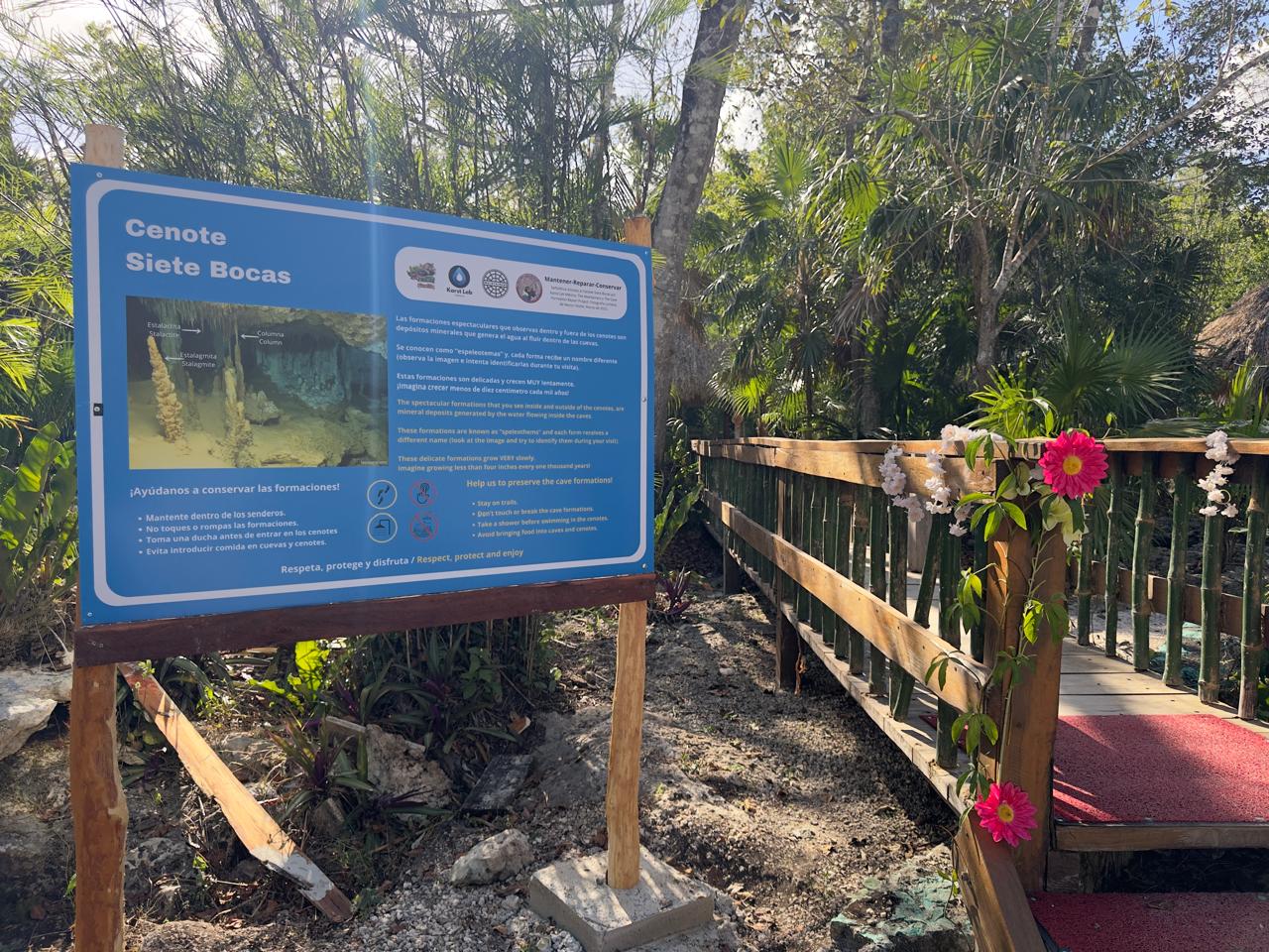 A large informational sign about "Cenote Siete Bocas" stands at the entrance of a wooden pathway surrounded by lush tropical vegetation. The sign includes images of cave formations and bilingual text in Spanish and English urging visitors to respect and protect the cave.